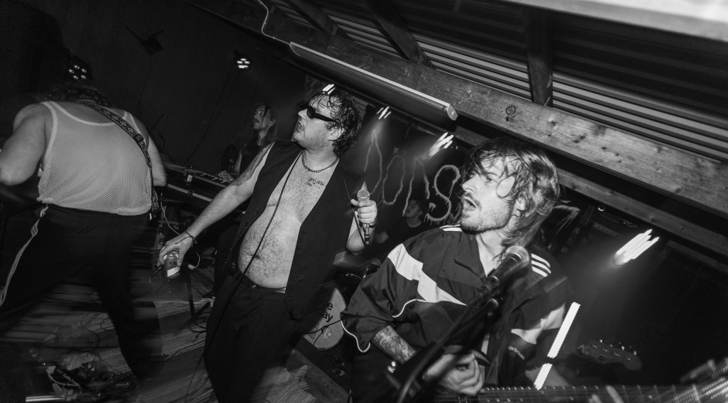 En överviktig och halvnaken musiker på scen med skinnväst och solglasögon.